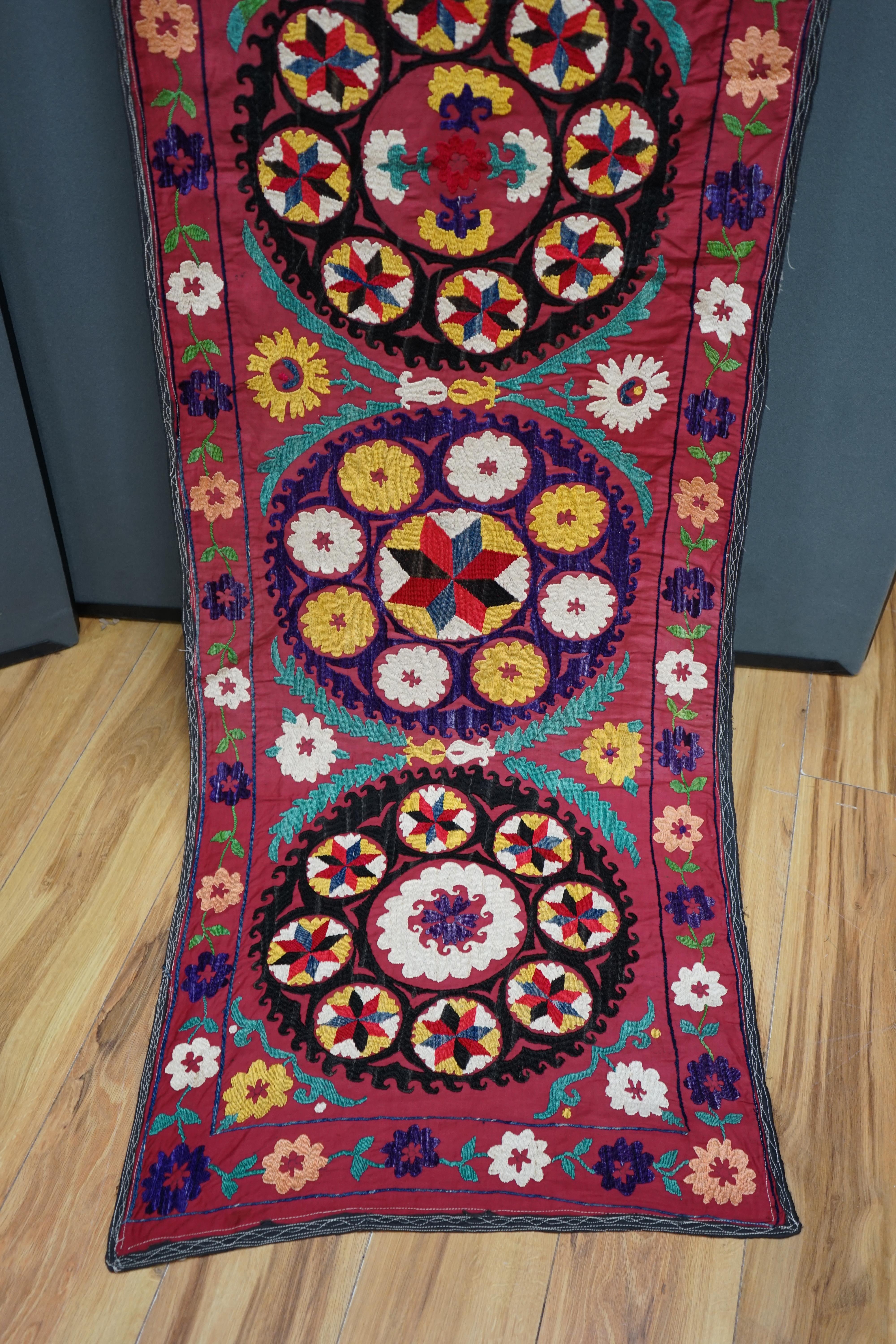 A 20th century Suzani wall hanging, possibly Uzbekistan, embroidered in bright polychrome silk threads into large symbols representing happiness, health and luck, on a burgundy cotton background, with a fine muslin backi